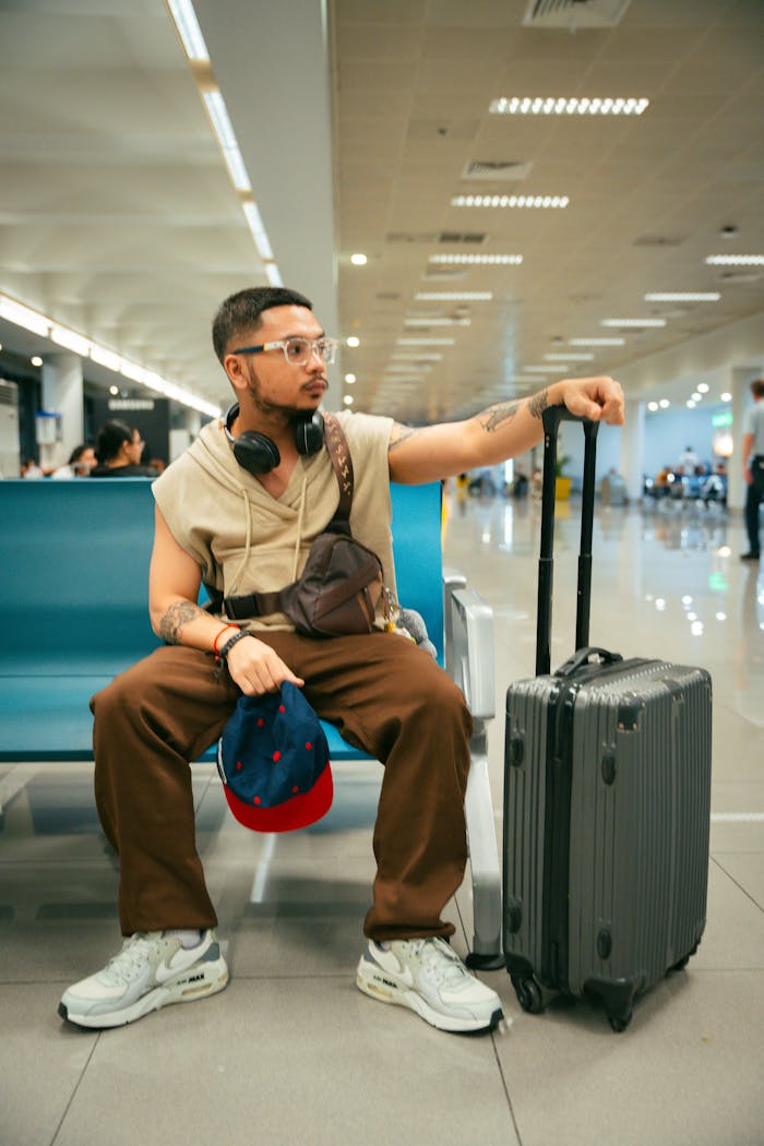 Stylish young man with suitcase seated in airport terminal, looking alert and prepared for travel.